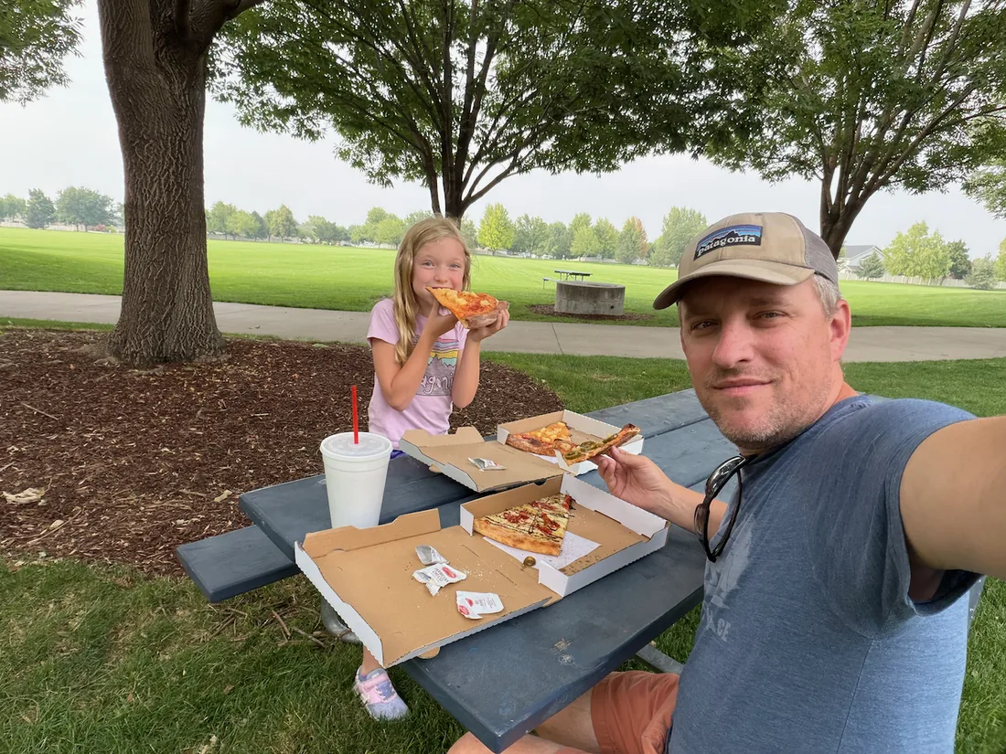 Phil and one of his kids eating pizza at a picnic table in a park.